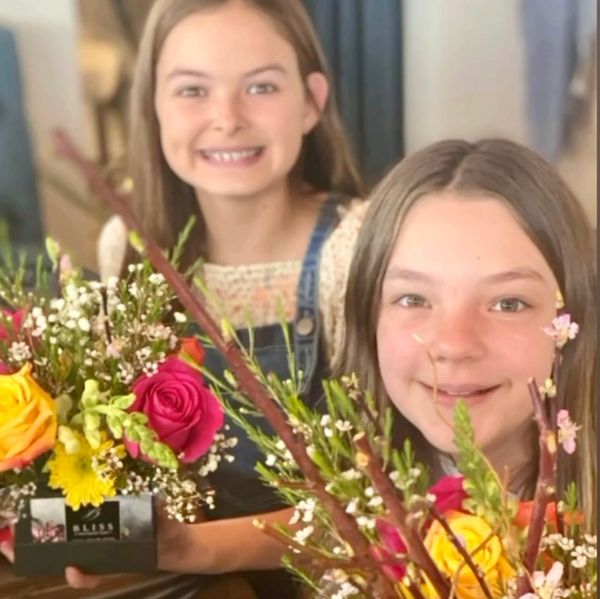 Two girls smiling behind vibrant floral arrangements.