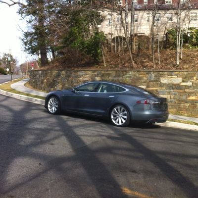 A dark gray Tesla parked on a suburban street near a stone wall.