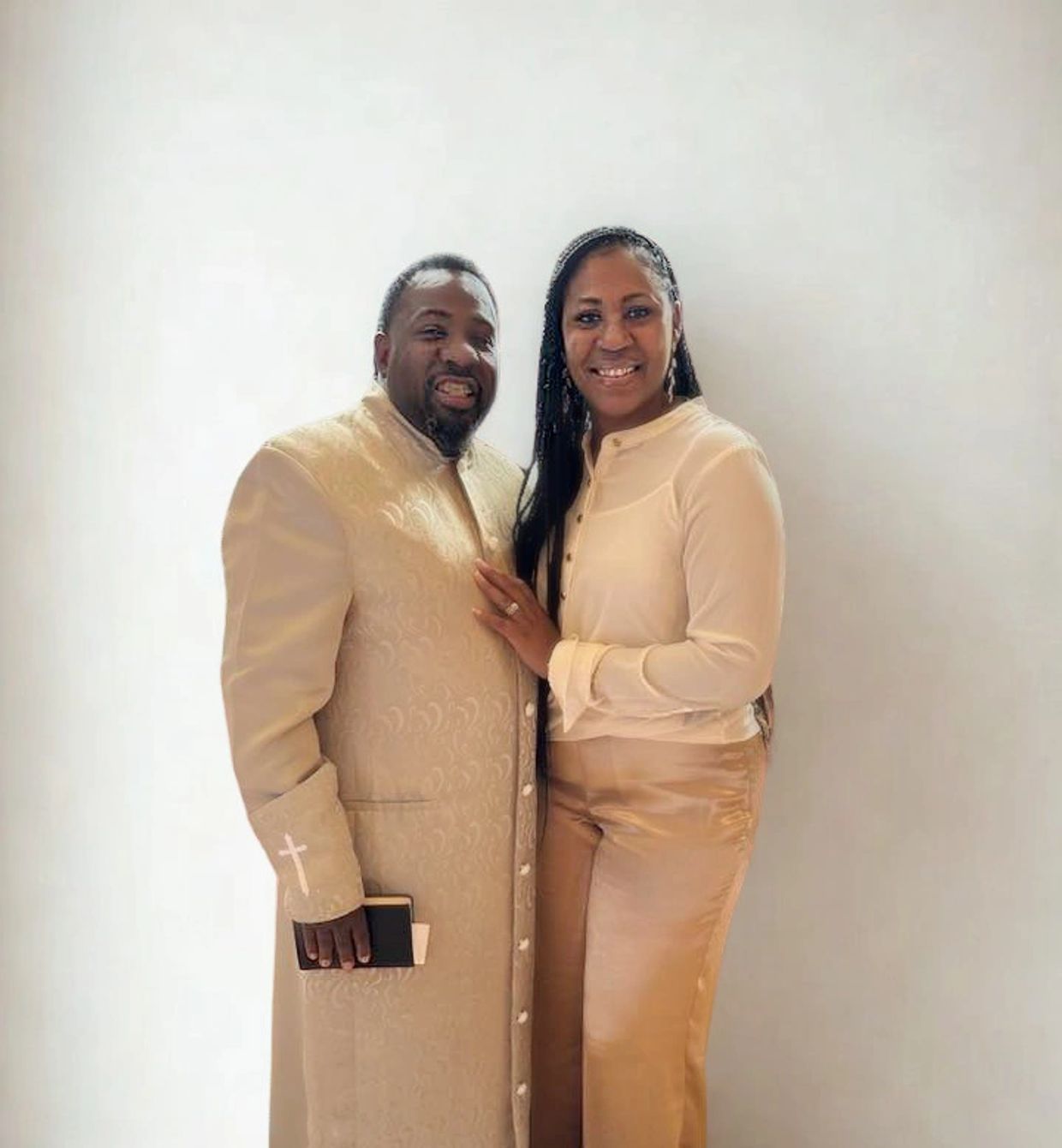 A couple dressed in elegant beige attire smiling against a plain background.