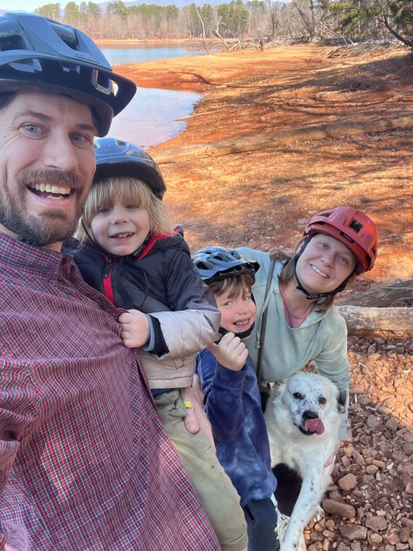 Family selfie with helmets near a lake on a sunny day.