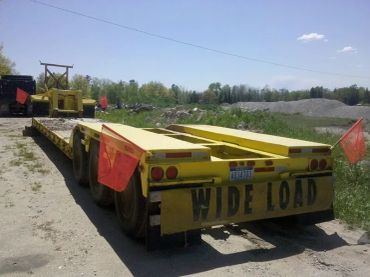 A yellow flatbed trailer labeled 'WIDE LOAD' with red flags on a dirt road.