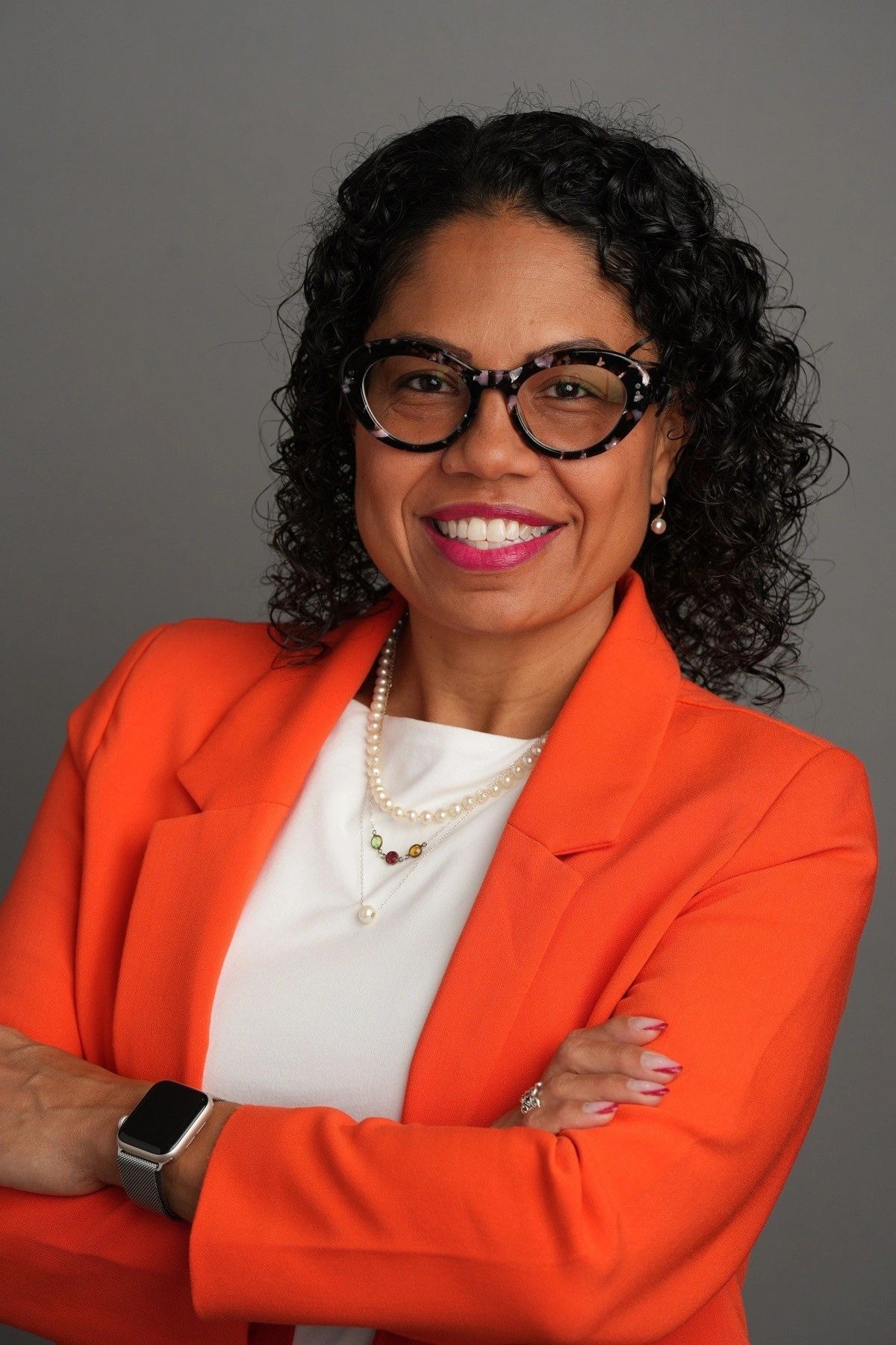 Confident woman in orange blazer smiling with arms crossed against a gray background.