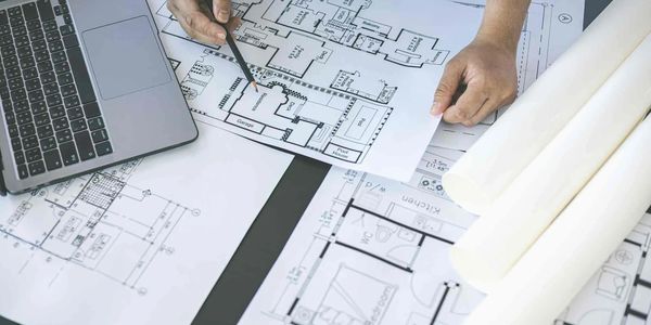 Manos señalando planos arquitectónicos al lado de una computadora portátil.