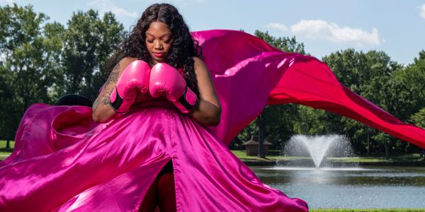 Woman in pink boxing gloves and flowing dress by a pond.