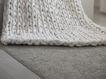 Close-up of a chunky knit white blanket on a gray carpet.