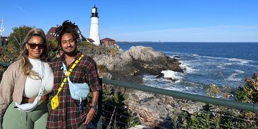 Couple posing by a rocky coastline with a lighthouse in the background on a sunny day.