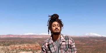 Man with dreadlocks in a plaid jacket under clear blue sky.