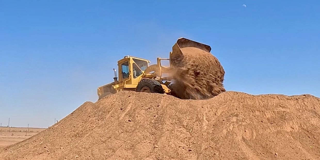 Yellow bulldozer dumping dirt on a large mound under clear blue sky.