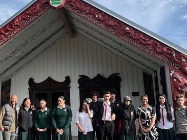 Group of students and adults standing in front of a traditional Maori meeting house.