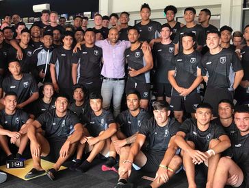Large sports team group photo indoors, most wearing matching black uniforms with one in casual.