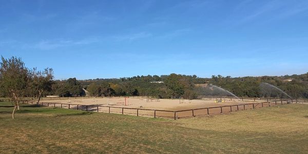 A fenced sandy area with sprinklers watering the ground under a clear blue sky.