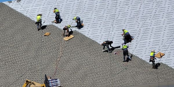 Workers installing shingles on a steep roof using safety harnesses and a lift.