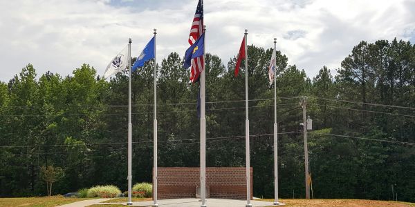 Five flags on flagpoles with a brick memorial and trees behind them.