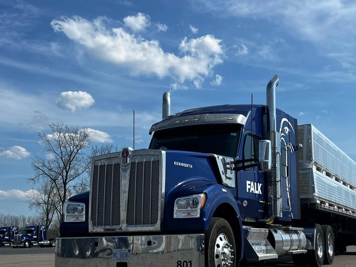 Blue Kenworth truck with FALK branding parked under a partly cloudy sky.