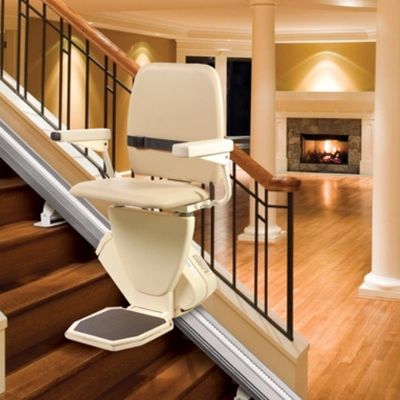 A beige stairlift installed on wooden stairs with a cozy fireplace in the background.