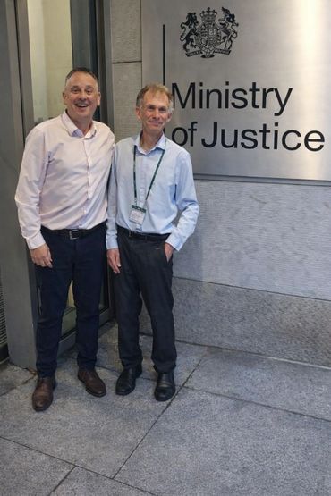 Two men standing and smiling next to a Ministry of Justice sign.