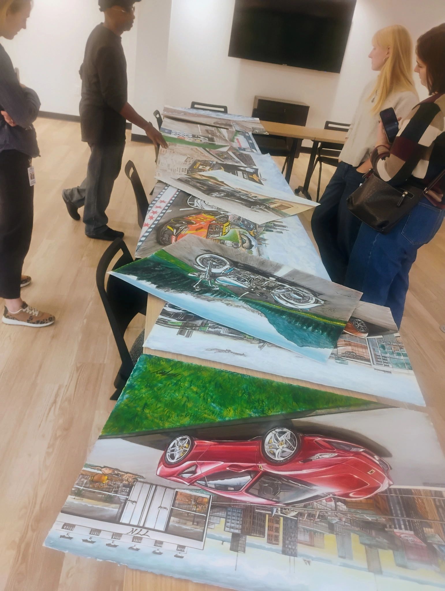 People examining automotive and landscape paintings on a table.