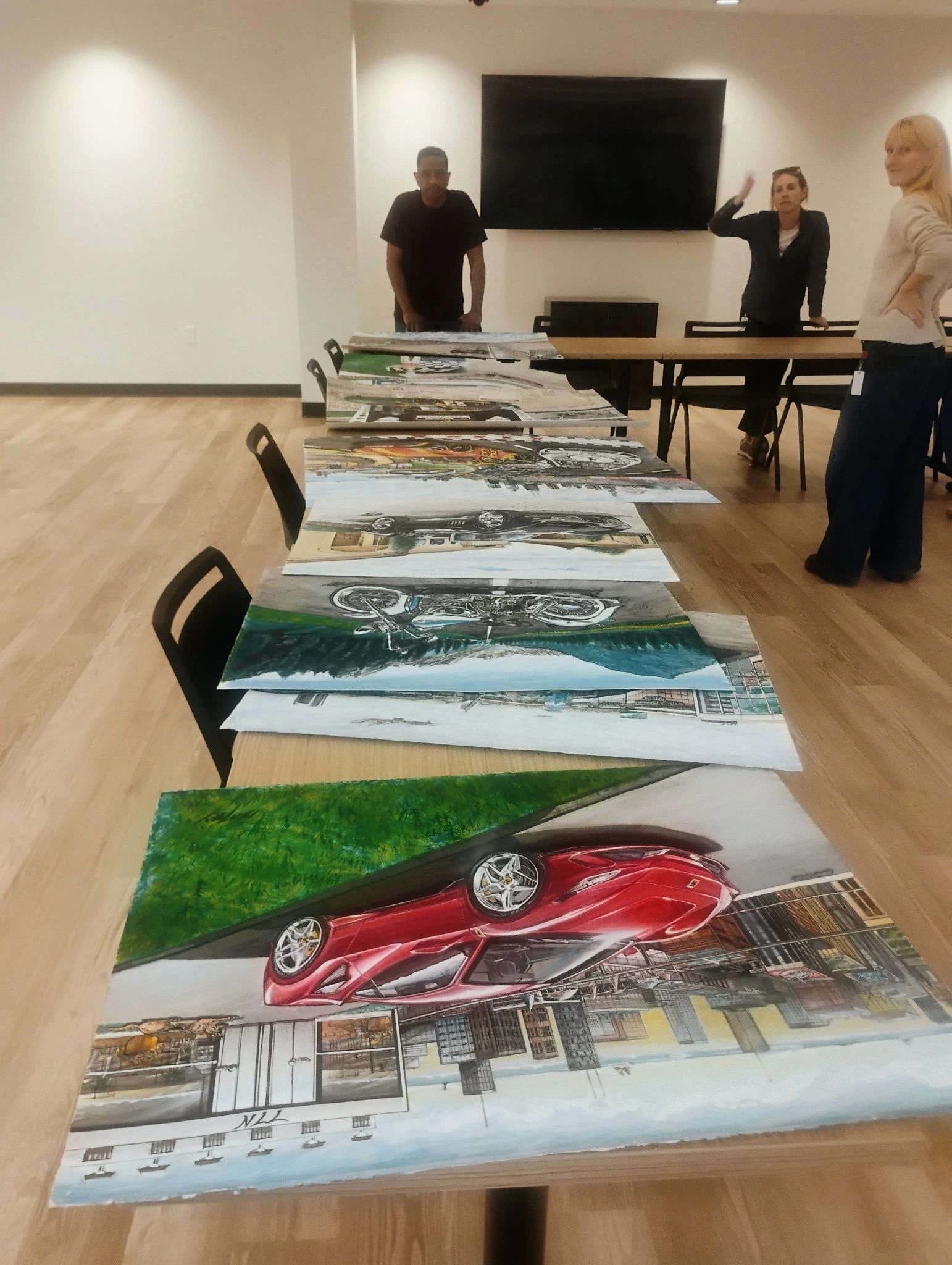 Artworks of cars displayed on tables with people in the background.