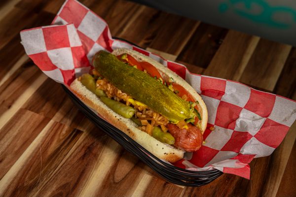 hot dog with pickle and toppings sits in a dish lined with red/white checkered paper 