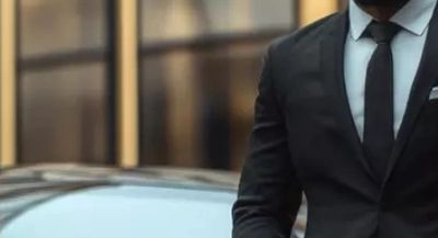 A man in a black suit standing in front of a car with a blurred background.