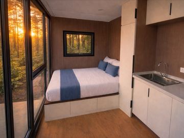Cozy bedroom with large windows and kitchenette in a cabin surrounded by forest.