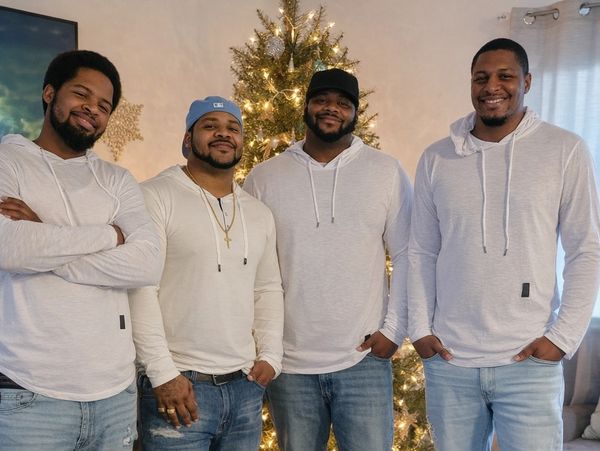 Four men in white hoodies and jeans smiling by a decorated Christmas tree.