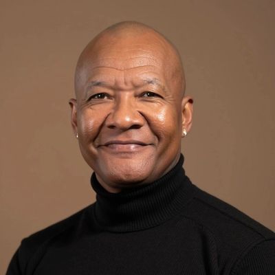 Smiling bald man wearing a black turtleneck against a brown background.