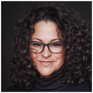 Smiling woman with curly hair and glasses against a dark background.