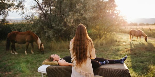 A woman receives outdoor therapy with horses grazing nearby at sunset.