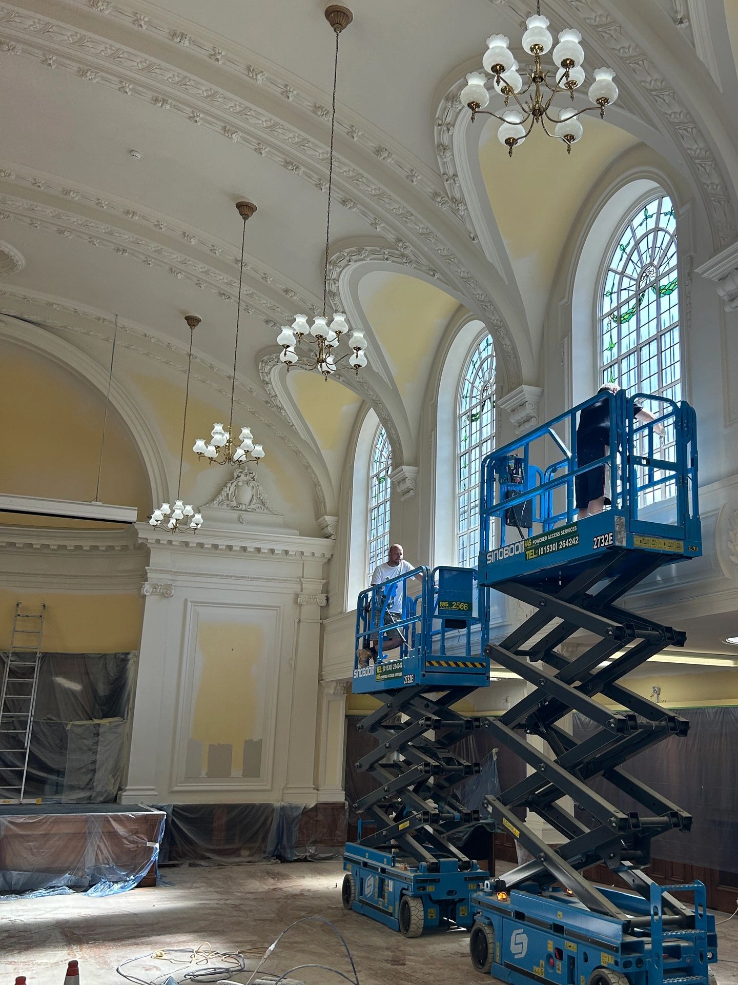 Workers on lifts restoring ornate windows in a grand hall.