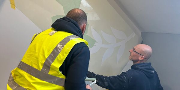 Two men painting a wall with leaf patterns indoors.