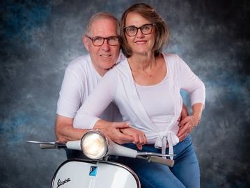 Elderly couple smiling on a vintage Vespa scooter against a studio backdrop.