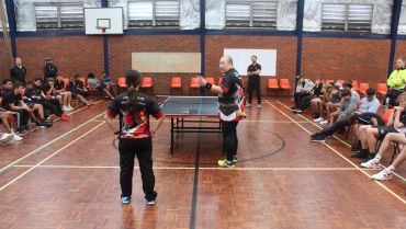 Two players facing each other at a table tennis match with audience seated on both sides.