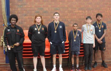 Six young athletes stand in a line wearing medals, posing indoors against a brick wall.