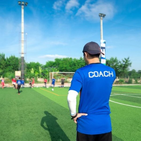 A coach watches young players practicing soccer on an outdoor field under a clear sky.