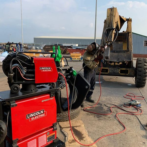 Worker using Lincoln Electric welding machine on heavy equipment outdoors.