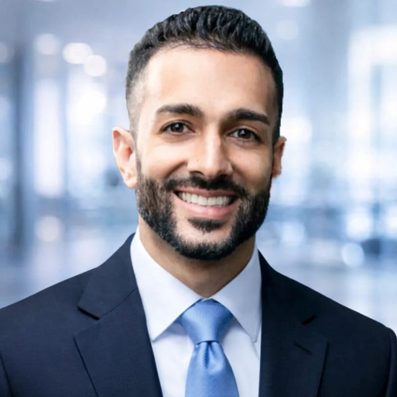 Smiling man in a suit with a blue tie against a blurred office background.