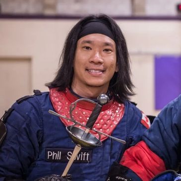 A smiling person in fencing gear holding a sword indoors.