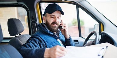 Man in a cap talking on phone and writing in a car.