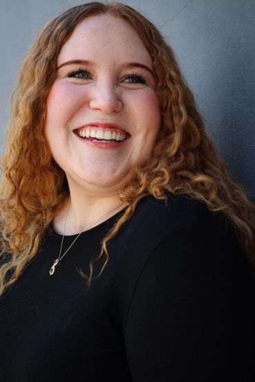 Smiling woman with curly red hair against a gray wall.