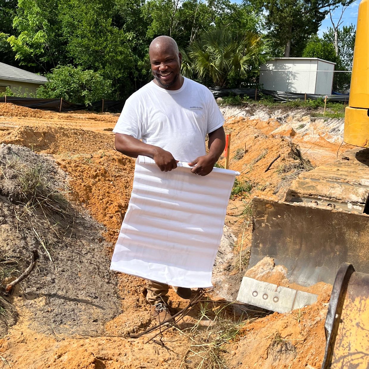 A man holding blueprints at a construction site with an excavator.