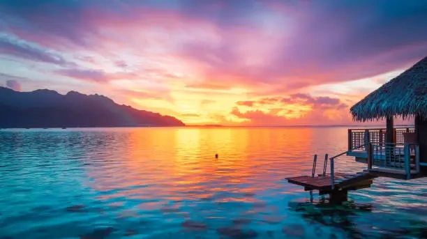 Sunset over serene water with mountains and a thatched hut.