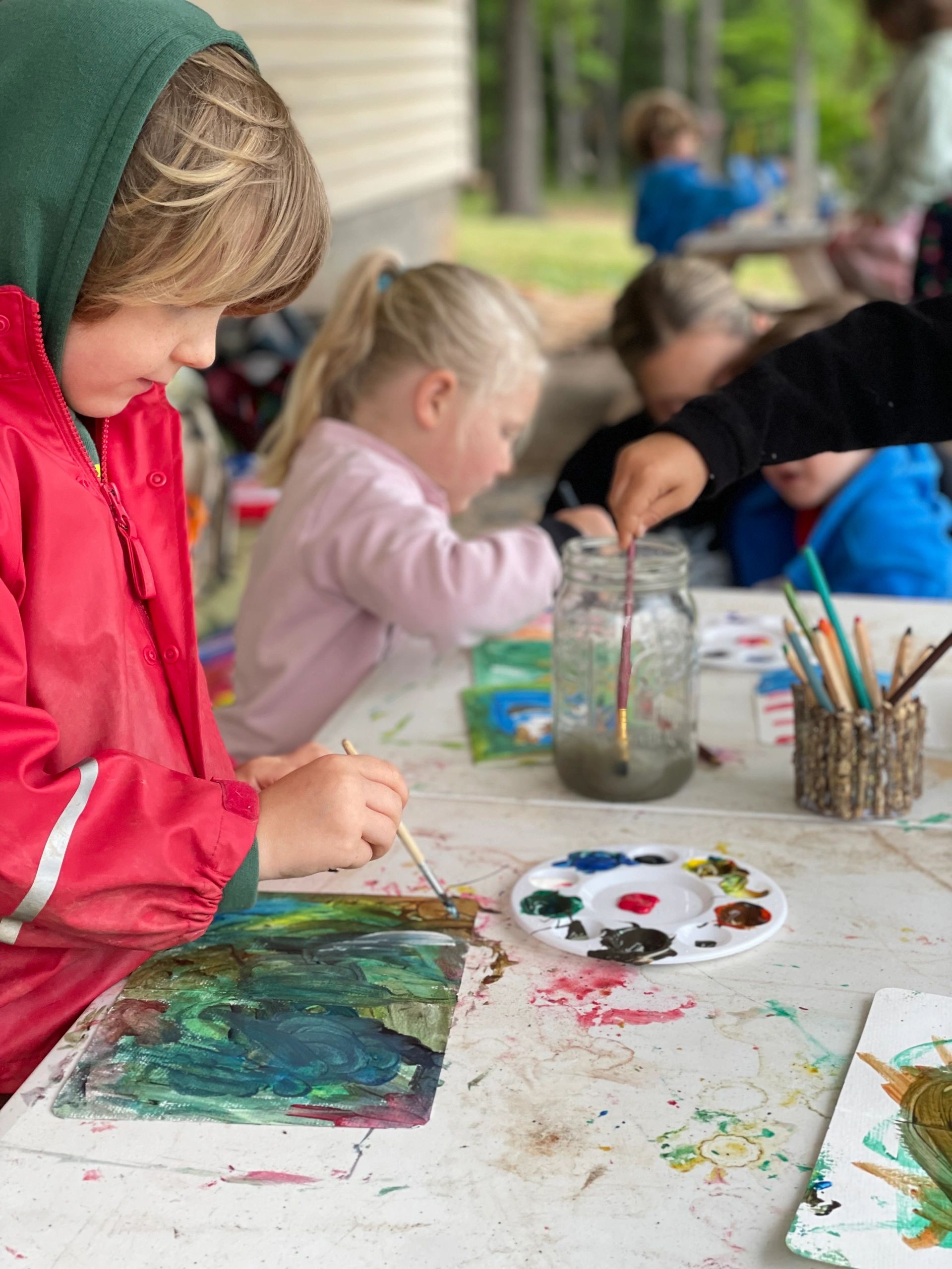 Children focused on painting at a messy art table outdoors.