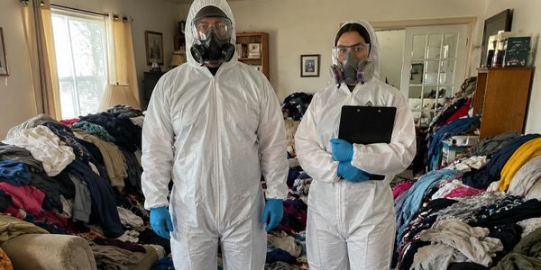 Two individuals in protective suits stand in a cluttered room filled with piles of clothes.