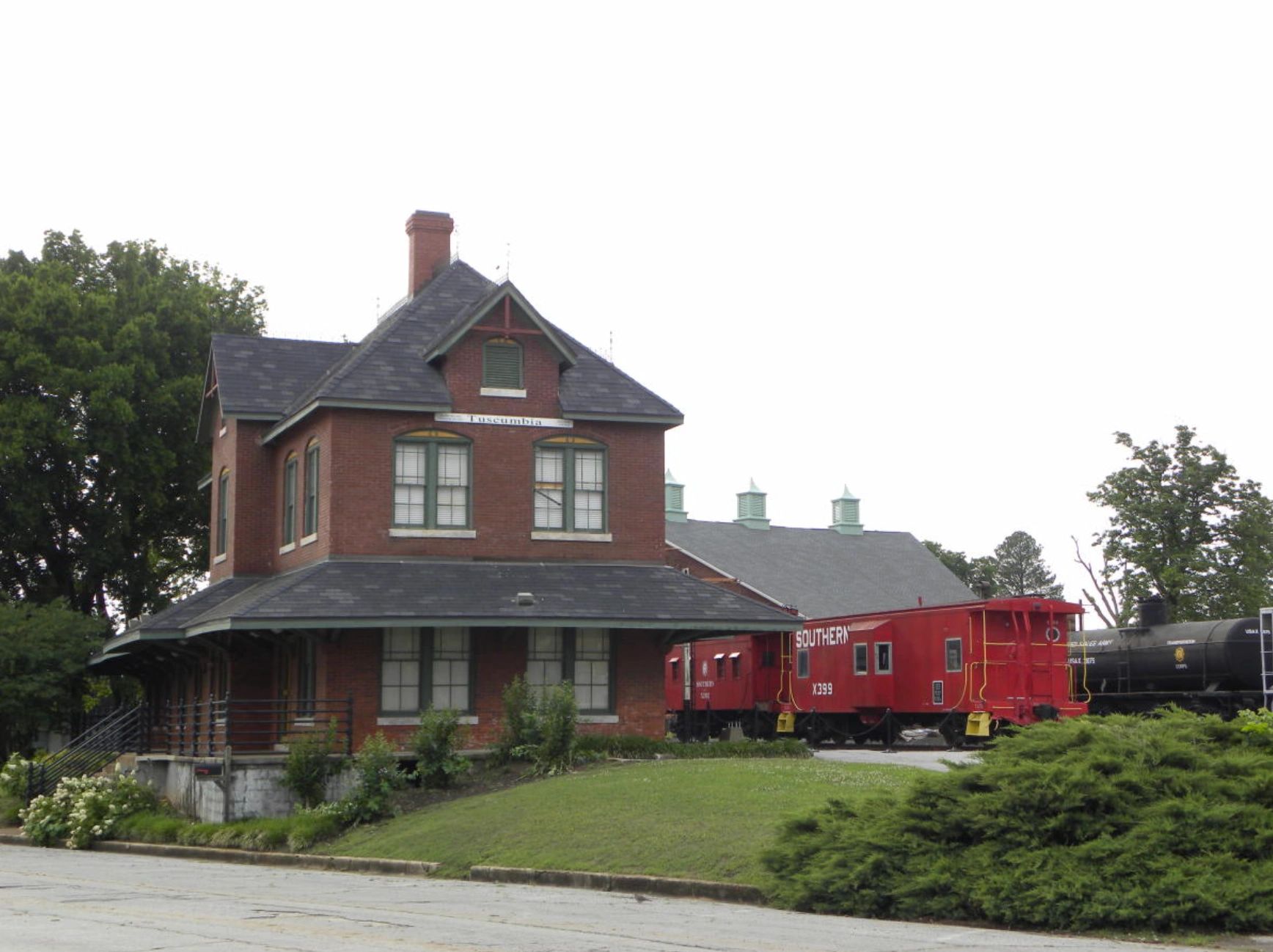 Historic brick train station with red train cars on tracks beside it.