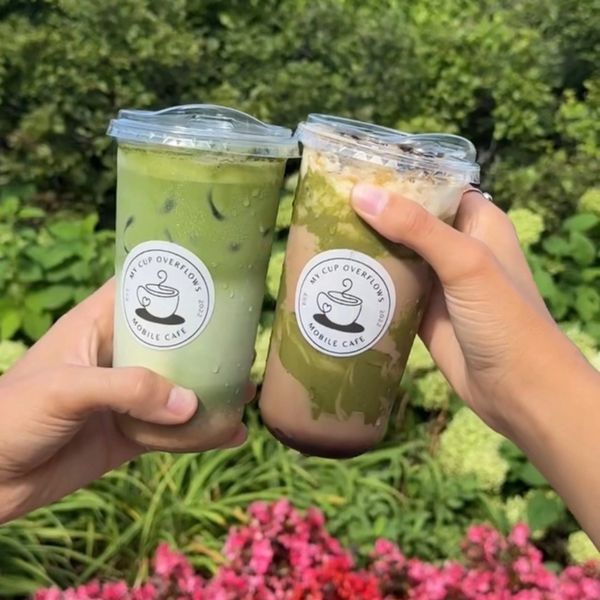 Two hands holding iced matcha drinks in clear cups outdoors.