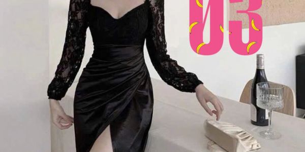 Elegant woman in a black lace dress with a thigh slit, standing by a table with a wine bottle and glass.