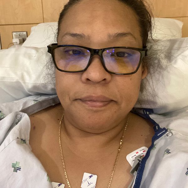Woman in hospital gown lying in bed, wearing glasses and a gold chain.