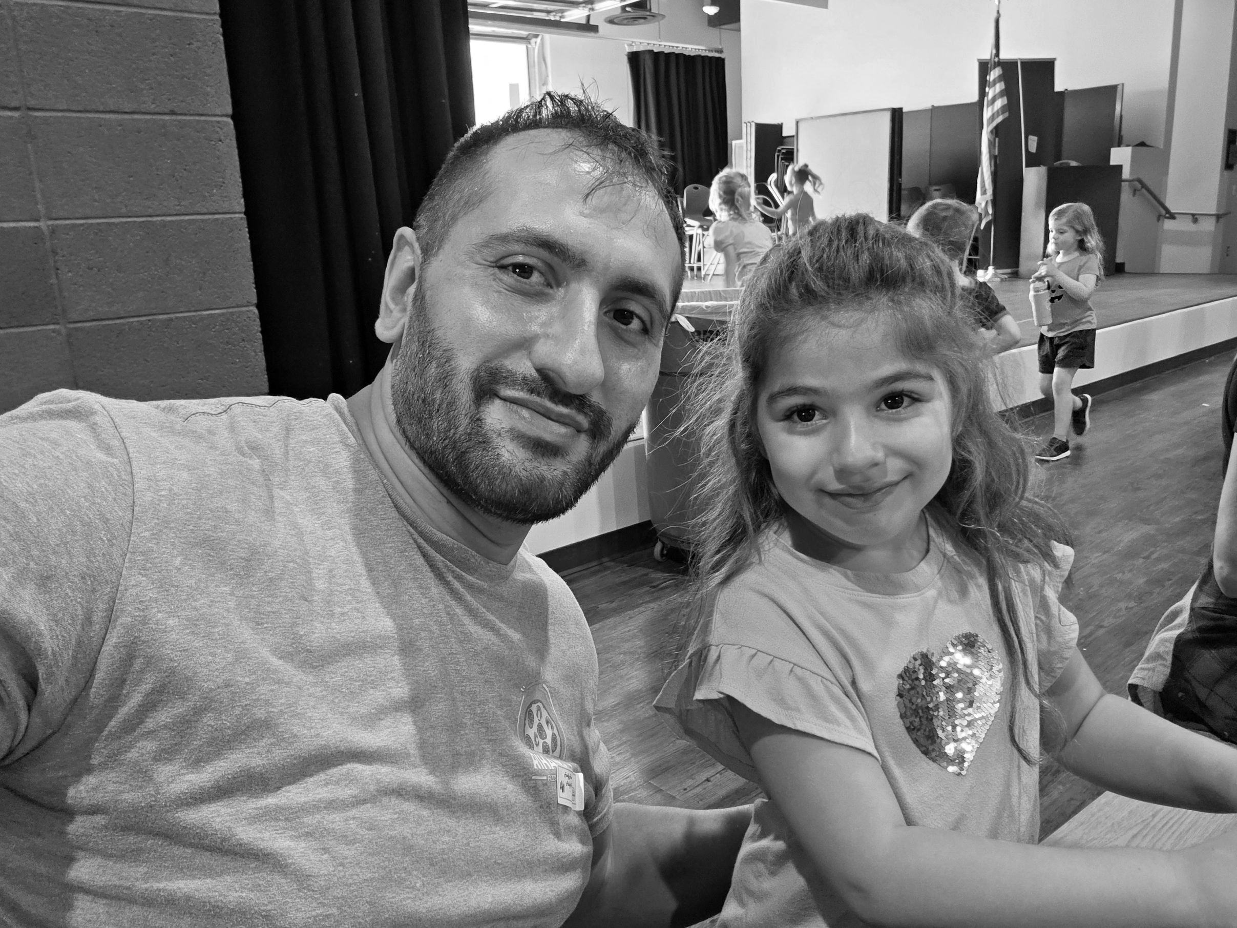 A man and a young girl smiling indoors, with children playing in the background.