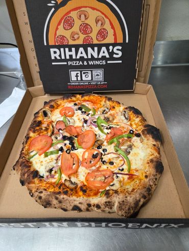 A freshly baked veggie pizza with tomatoes, olives, and peppers in a Rihana's pizza box.